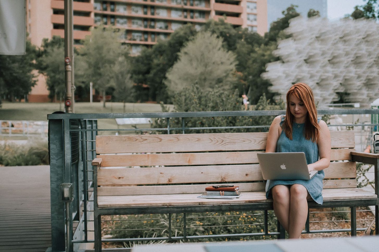 A woman sits on a park bench while working on a laptop computer.