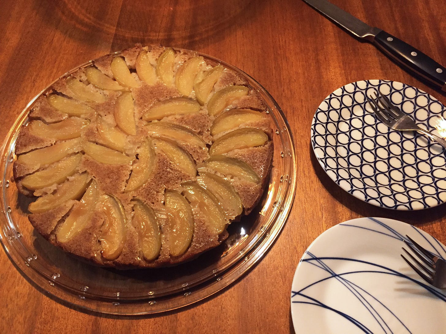 on a cake plate, a craggy apple cake with a spiral of apple slices. Next to it are two dessert plates with forks on them, and above them is a large knife to cut the cake.