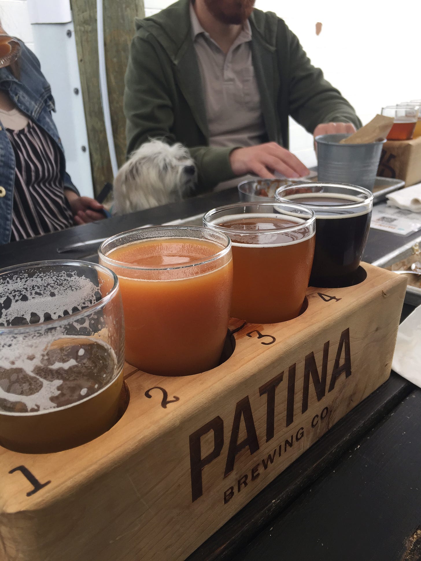 A flight of a variety of different-coloured beers in a wooden block that reads "Patina Brewing Co." In the background, Jesse and Krystyna are partially visible with their white dog, Wally, sitting between them.