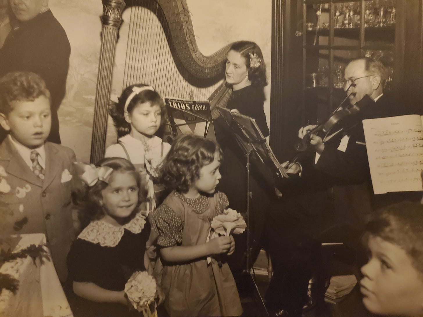 A sepia toned photograph of five small children in dress clothes in front of a harp player and violinist. 