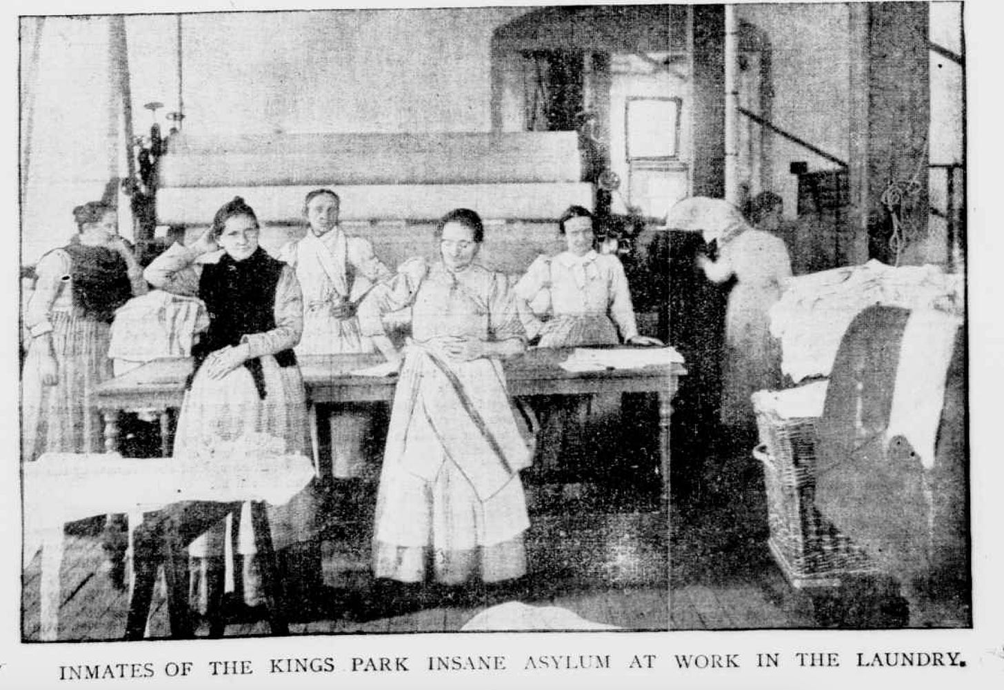 Black and white newspaper photo; there are women standing around a large laundry press. Caption reads: INSANE WOMEN AT WORK IN ONE OF TIIE SEWING CORRIDORS.