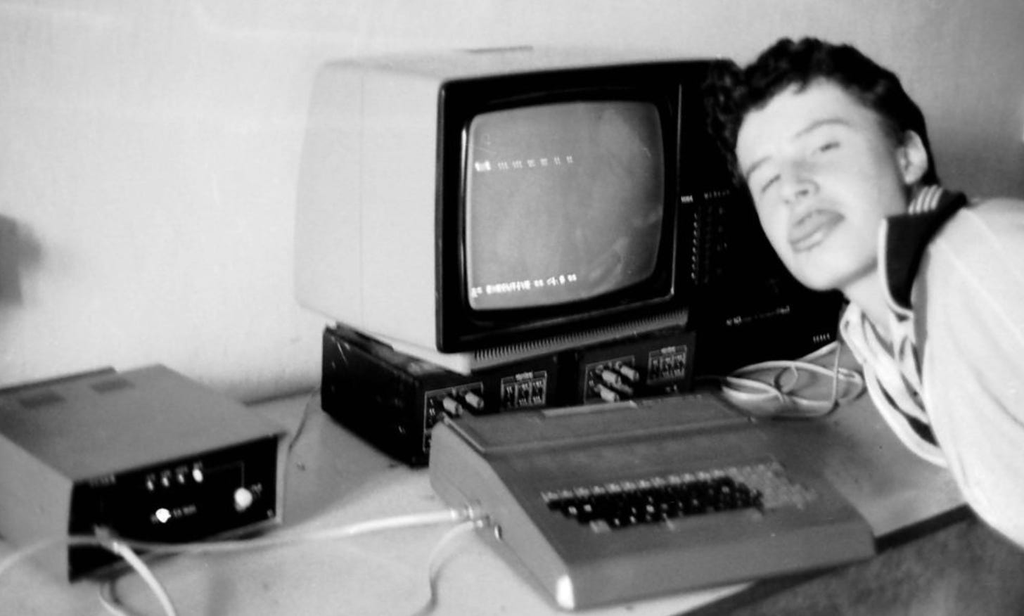 A teenager sticks his tongue out at the camera, sitting in front of a clunky 1980s computer with CRT screen.