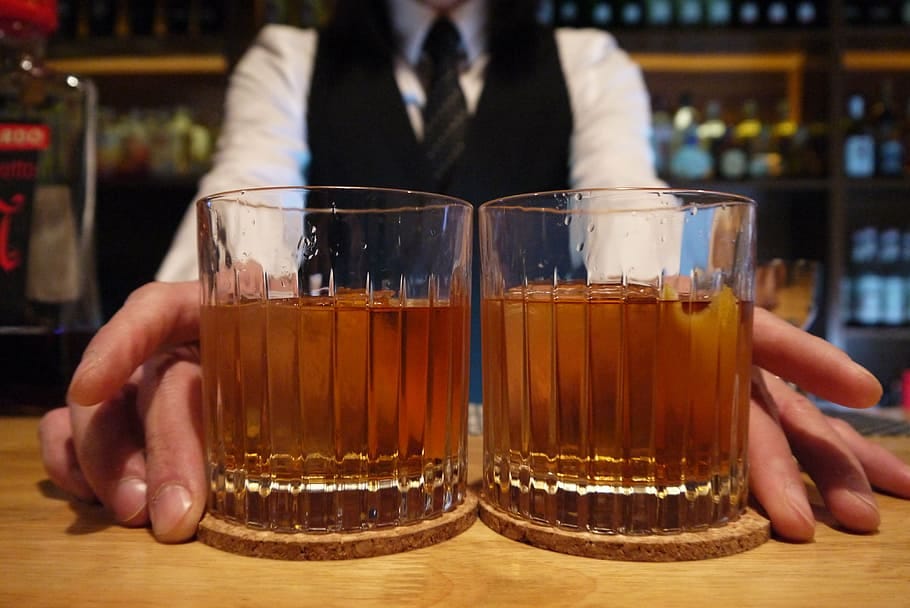 A faceless bartender presents two identical drinks in tumbler glasses.