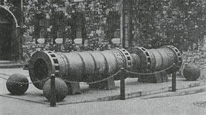 black and white image of a super canon flanked at all corners with canon balls, within a chained rectangular outdoor display area. Stone wall is in the background