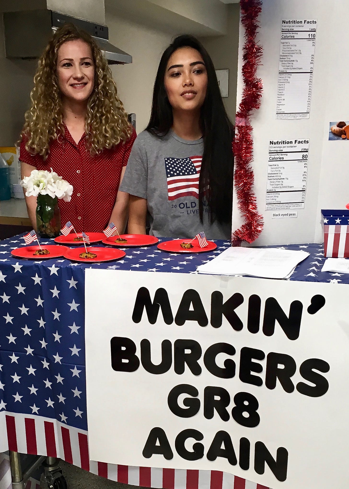 Two young women stand at a red-white-and-blue–themed table with veggie burger samples and the slogan Makin' Burgers Gr8 Again