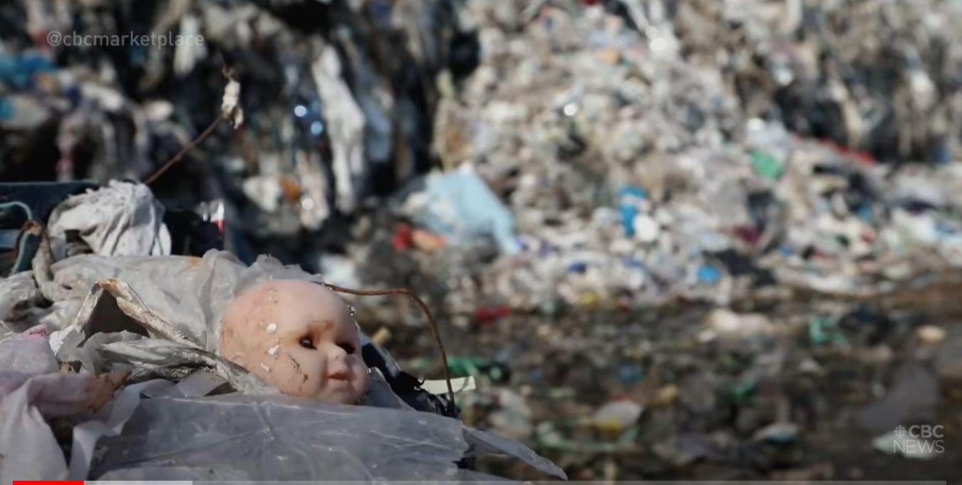 A bald doll head with one eye closed, sitting in a landfill.