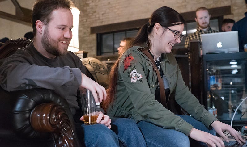 Two people sit together on a couch, drinking beer and playing a game on a laptop