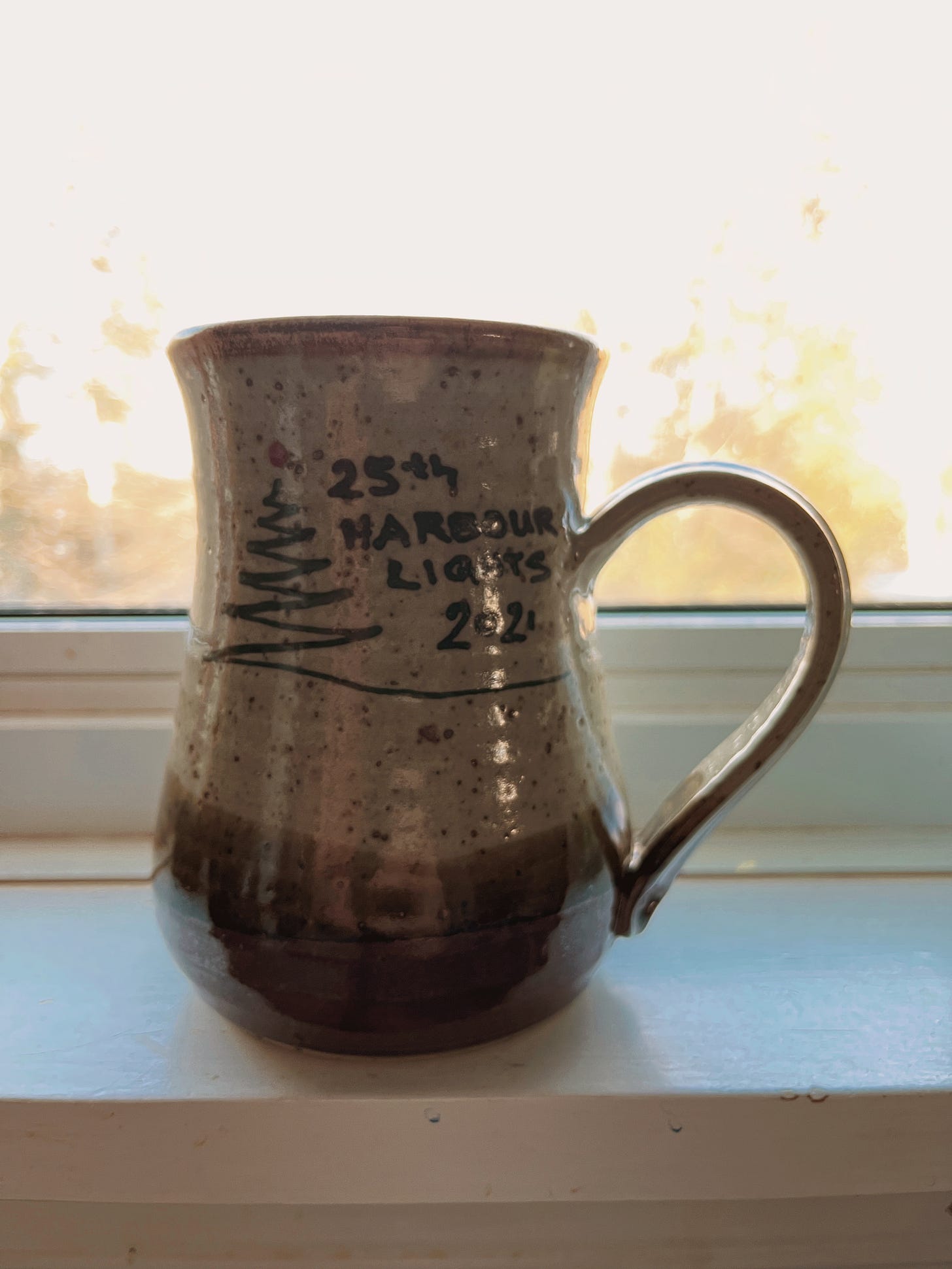 Tan and brown spotted pottery mug in front of a bright window. The words "25th Harbour Lights 2021" are written on the front of the mug.