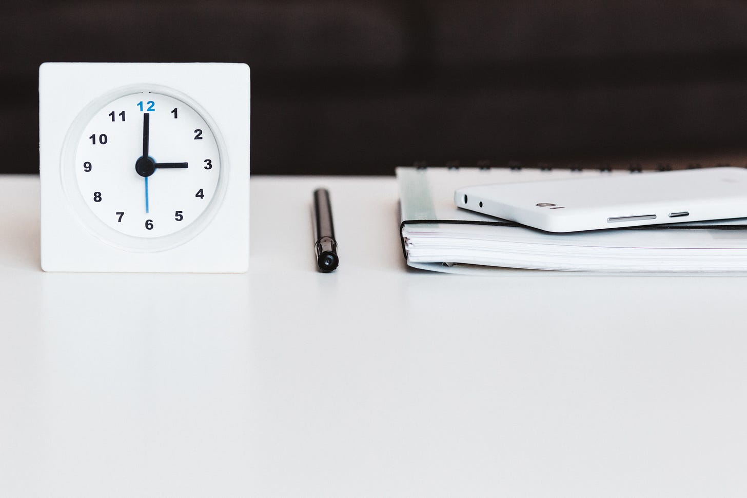 White clock on a desk with a black pen, notebook and a white phone