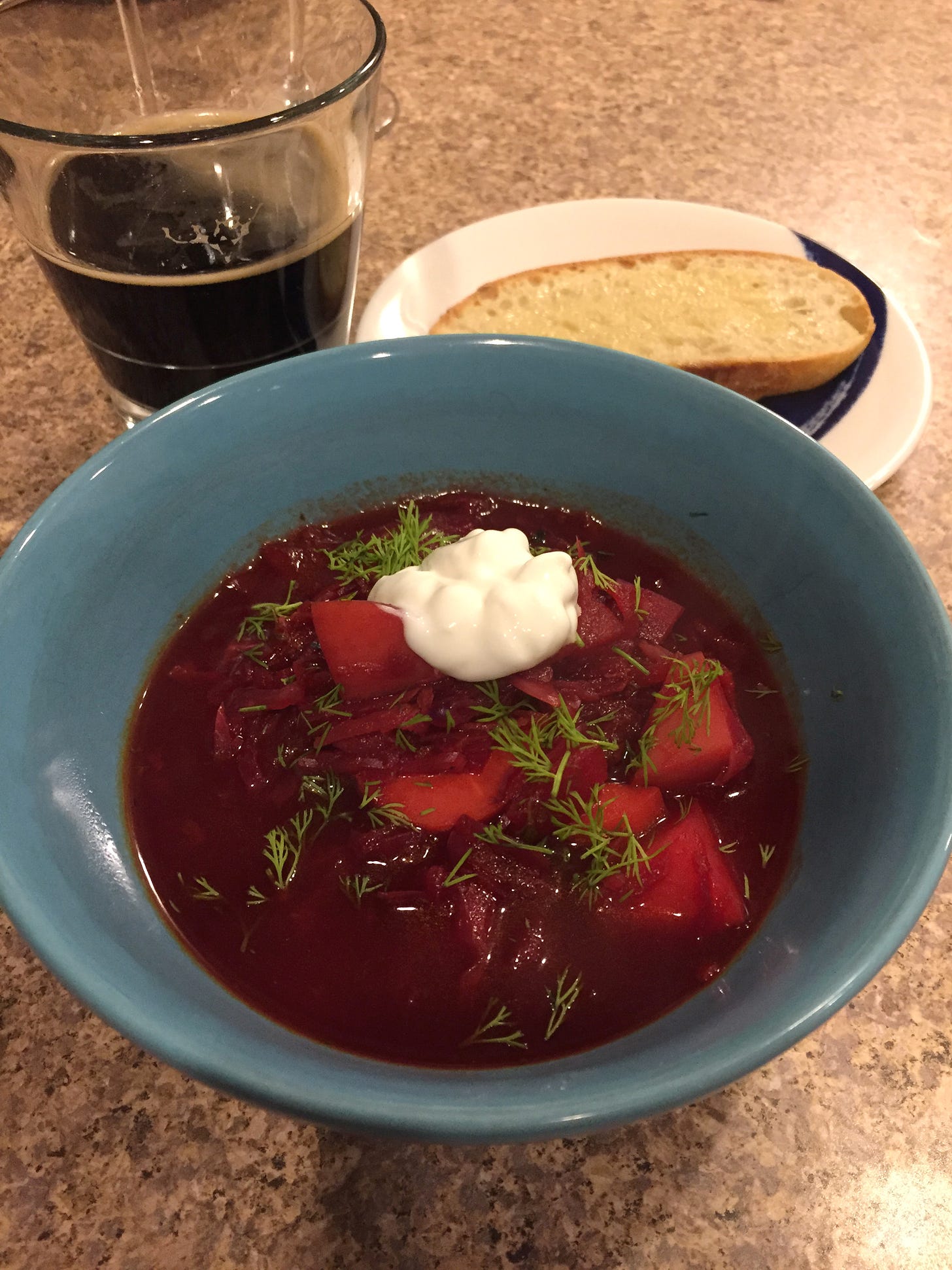 A blue bowl of borscht in the foreground, topped with chopped dill and a dollop of sour cream. Behind it is a small plate with a piece of sourdough toast, and a glass of dark beer.