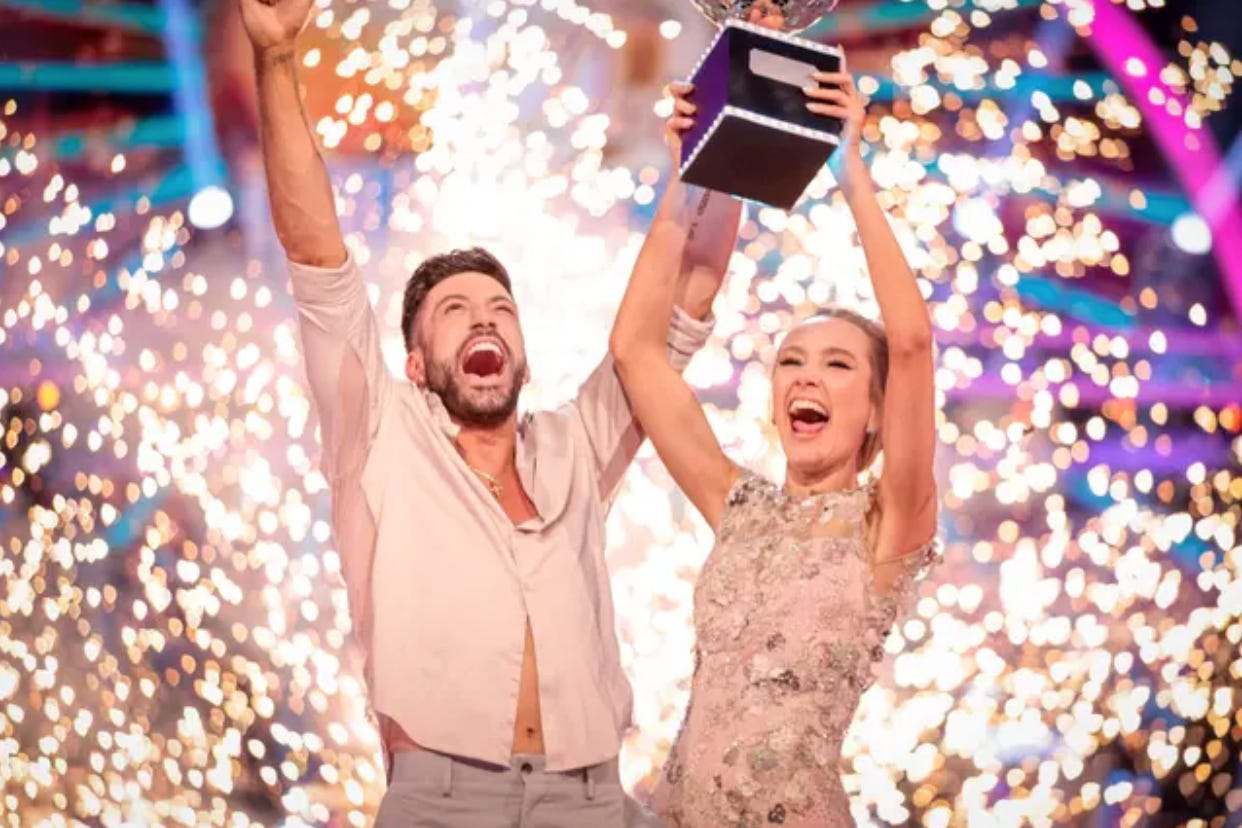 Giovanni Pernice, an Italian man, wearing a pale pink button shirt, is standing next to Rose Ayling-Ellis, a white, Deaf woman wearing a pale pink embroidered dress. They are laughing and holding up the Strictly Come Dancing Glitter Ball. Fireworks are exploding behind them.