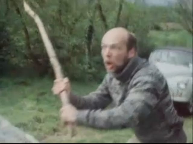 Man in a roll neck sweater wields a large stick with rage on his face