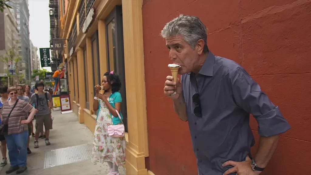 A still from Roadrunner. Anthony Bourdain eats an ice cream cone in front of a dark red wall.