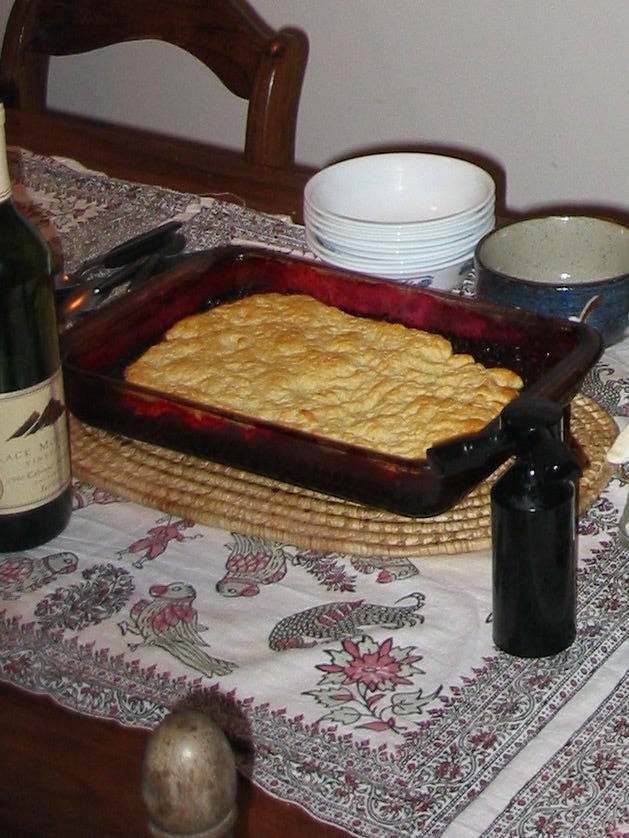 A large pan of duff sits on a wooden table covered with a tablecloth. Scattered around it are a stack of bowls, a bottle of wine, and a pepper grinder 