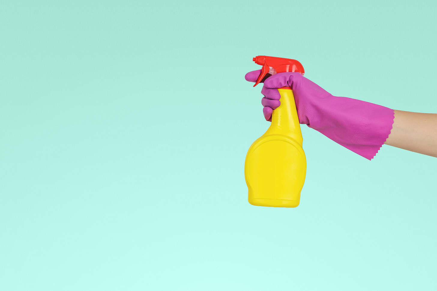 A hand wearing a purple rubber glove and holding a yellow and red spray bottle against a teal background