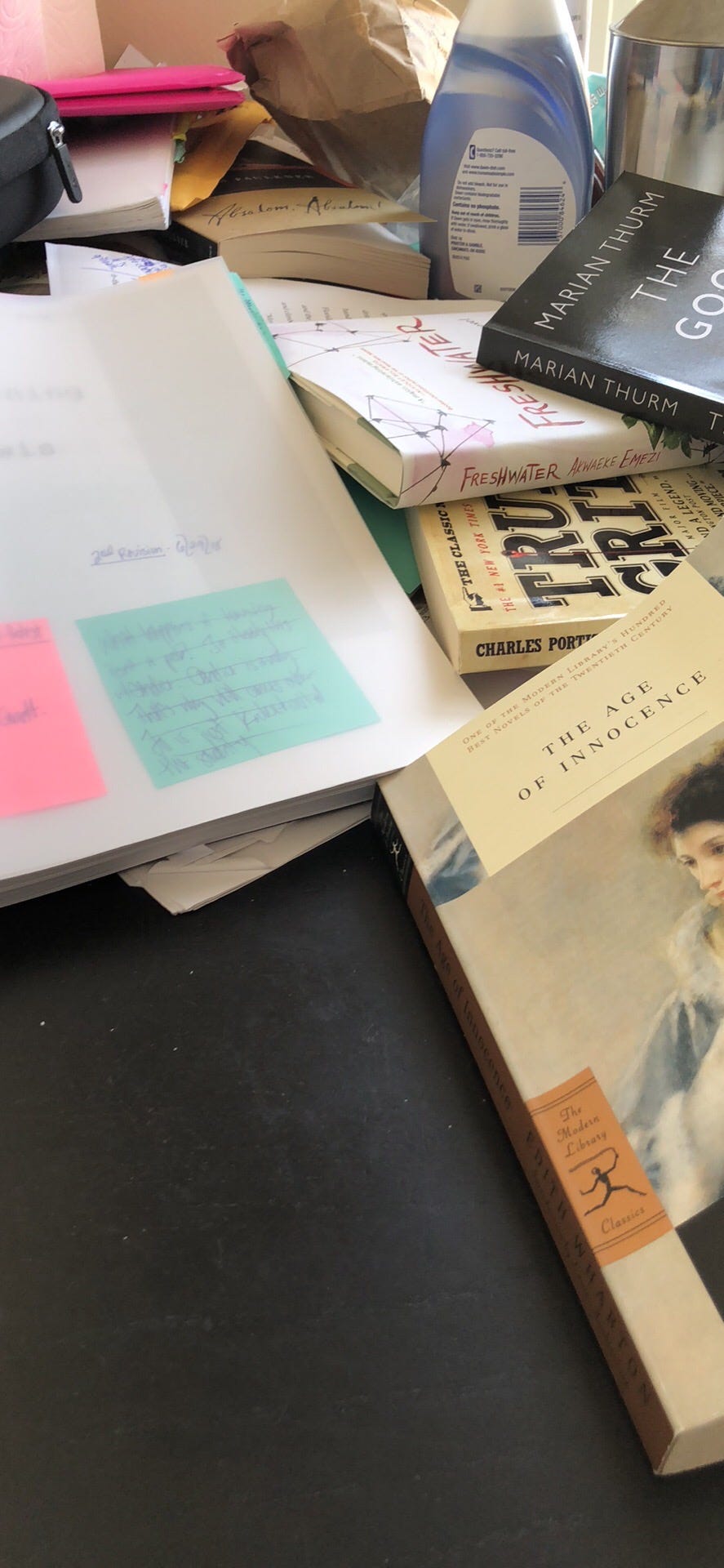 A photo of Kat’s messy writing desk. A print-out of her thesis novel is sitting next to True Grit by Charles Portis, The Age of Innocence by Edith Wharton, Freshwater by Akwaeke Emezi, and The Good Life by Marian Thurm.