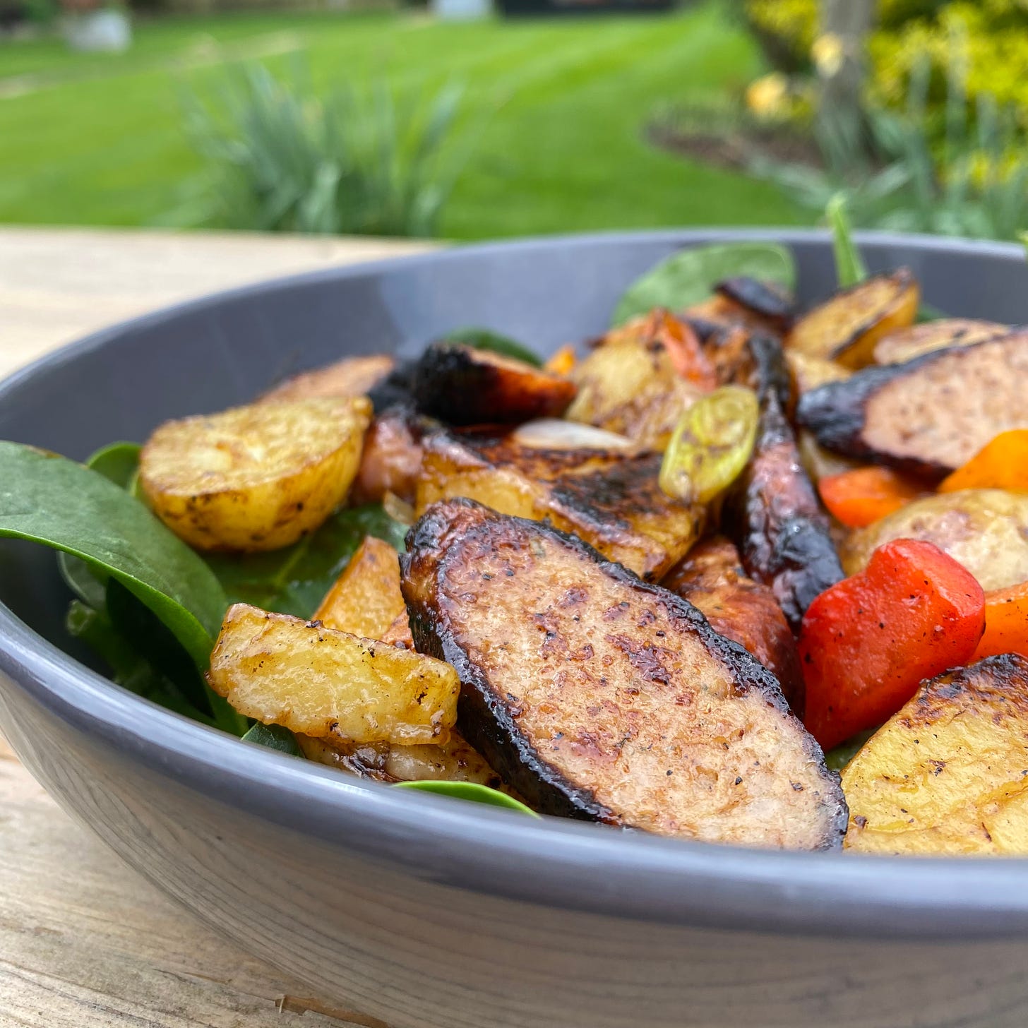Bowl filled with sliced sausages, peppers, potatoes and salad leaves on a table outside with garden in the backgroud