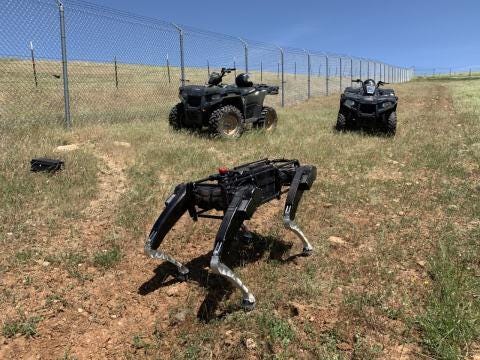 A black and silvery 4-legged metallic robot dog is walking across dry grass and dirt somewhere in the southwestern US. Two black ATVs are in the background near chain link fencing.