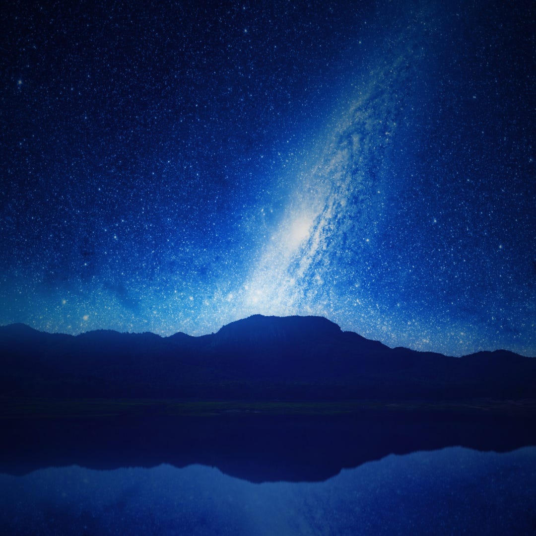 The Milky Way appears as spiral galaxy behind a far away mountain range at night. The stars are brilliantly bright and light up the sky around it in shades of cool blue.