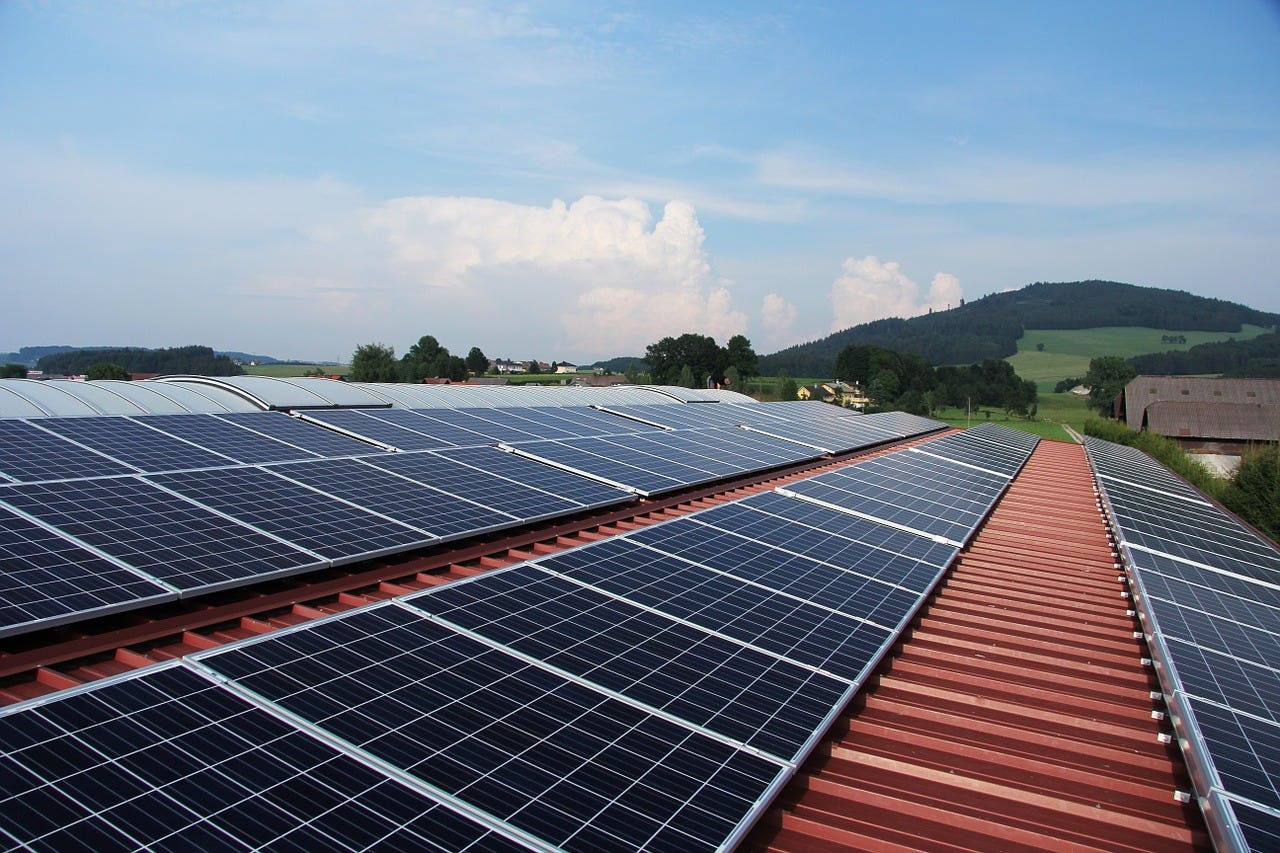 An array of solar panels on a red roof with green hills and a blue sky in the background.