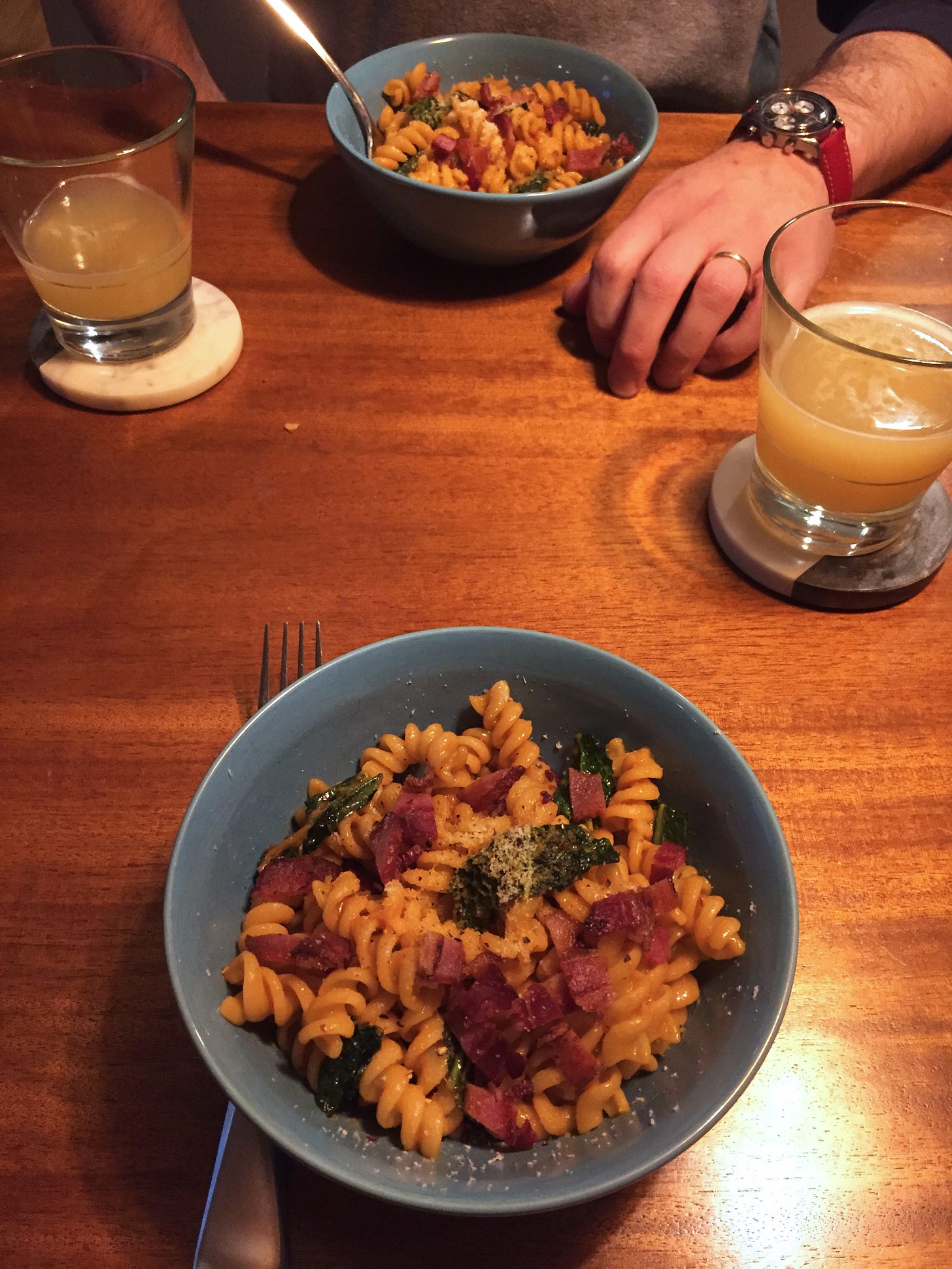 Across from each other on a table are two blue bowls filled with fusilli in vodka sauce, with pieces of kale throughout. A crumble of bacon is sprinkled across the top of each dish. On coasters are two partially-empty glasses of beer.