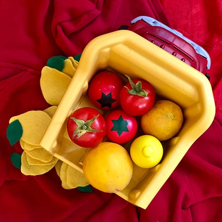 A still photo of a red and yellow plastic dump truck filled with two real tomatoes and two wooden tomatoes, and two real lemons and a wooden lemon, with felt lemons surrounding the truck.
