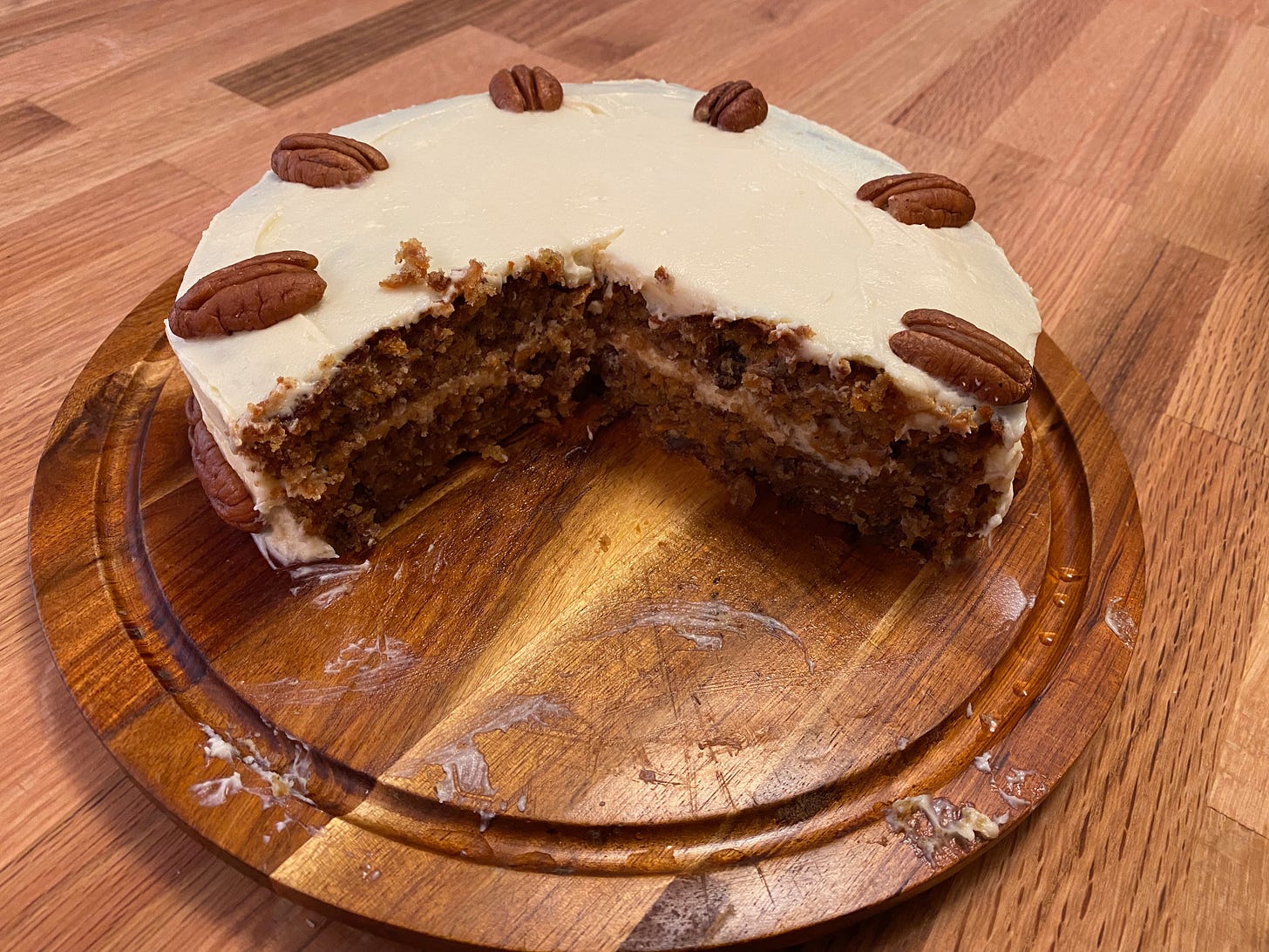 A two-layer carrot cake sits on a wooden platter. A large slice is cut out of it, revealing the layers inside. It’s frosted with white cream cheese frosting, and several pecan halves are arranged in a circle around the top.