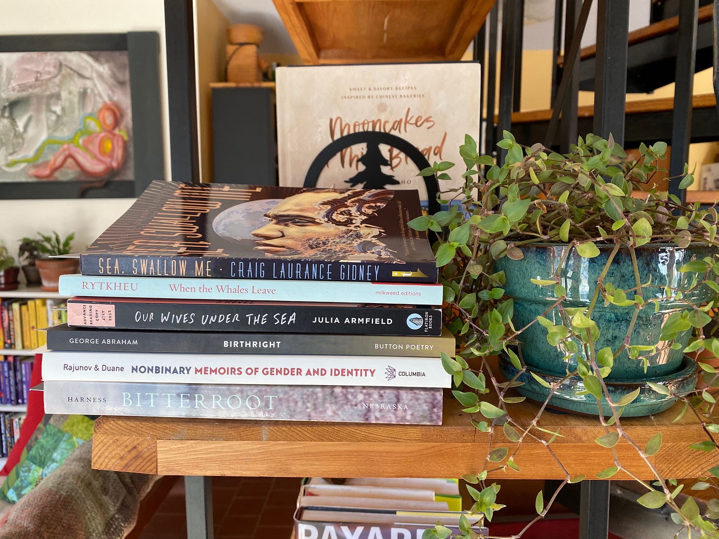 A stack of books on the end of a wooden bookshelf next to a plant with trailing green leaves in a small green pot. There is a cookbook standing upright on the shelf behind the book stack, and a painting on the wall and another bookcase visible in the background. The books are: Bitterroot, Nonbinary: Memoirs of Gender and Identity, Birthright, Our Wives Under the Sea, When the Whales Leave, and Sea, Swallow Me.