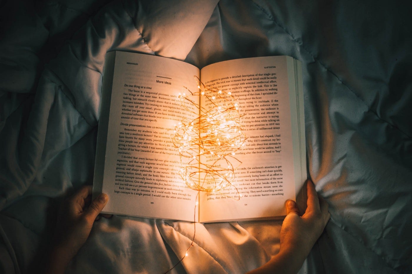 An open book being held by two hands on a bed cover. Small lights on a wire, like Christmas lights, are resting in the open crevice and lit, illuminating the words on the pages.