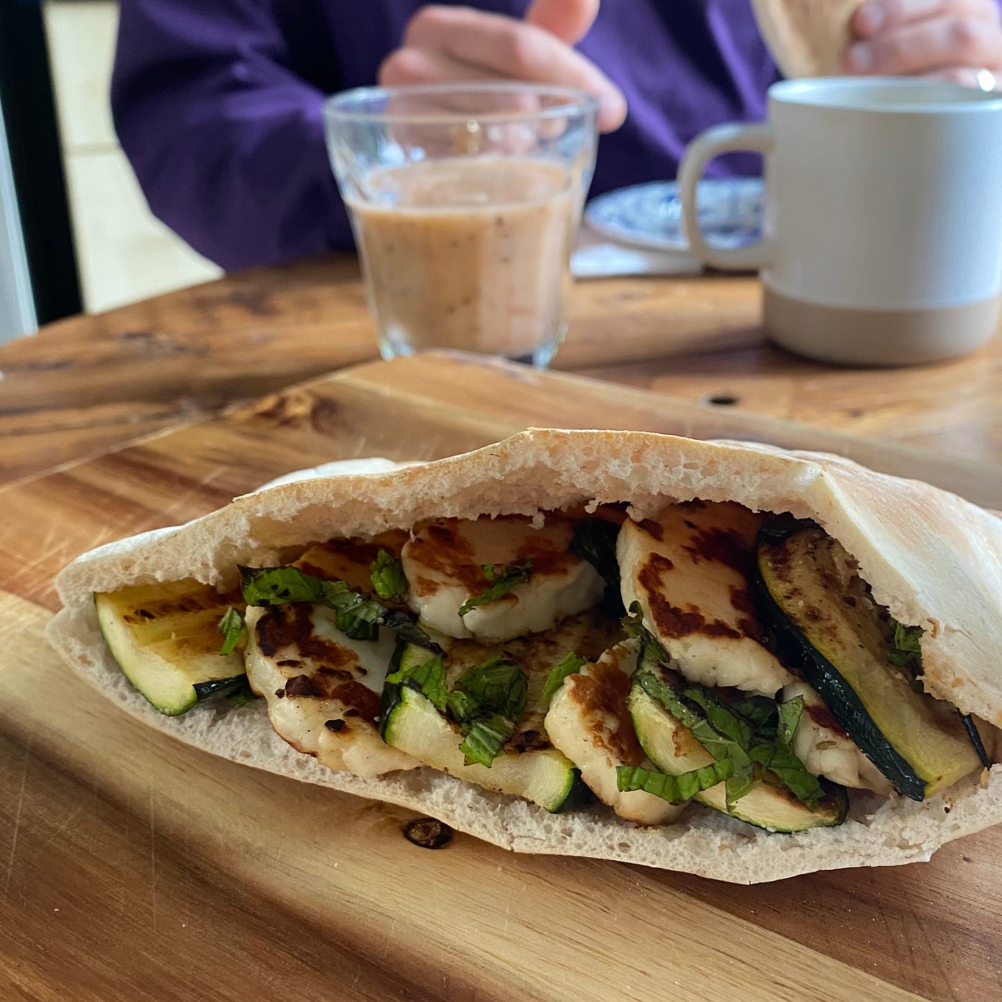Open pitta pocket filled with halloumi, sliced courgette and chopped mint. Pitta is on a wooden board, a cup, glass and a figure in the background