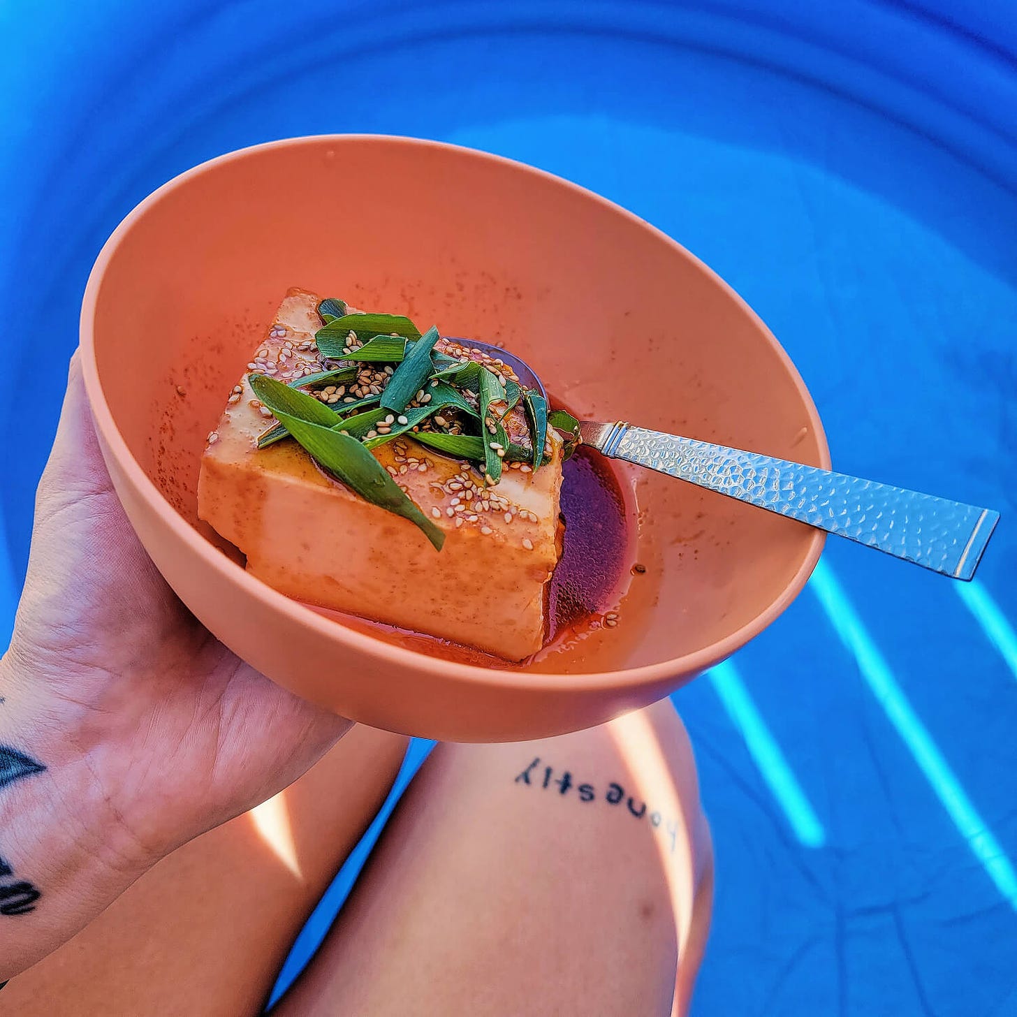 a block of silken tofu doused in a red gochujang sauce, topped with sliced green onions and toasted sesame seeds. grace is enjoying this in a plastic bowl with their feet dipped into a small inflatable backyard pool.