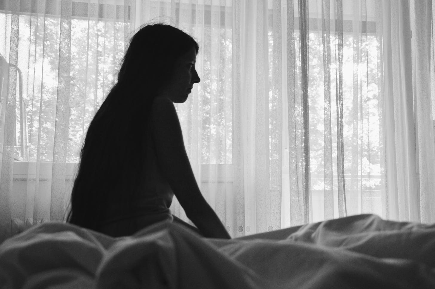 A woman sits in shadow on a bed. She is alone in a white room.