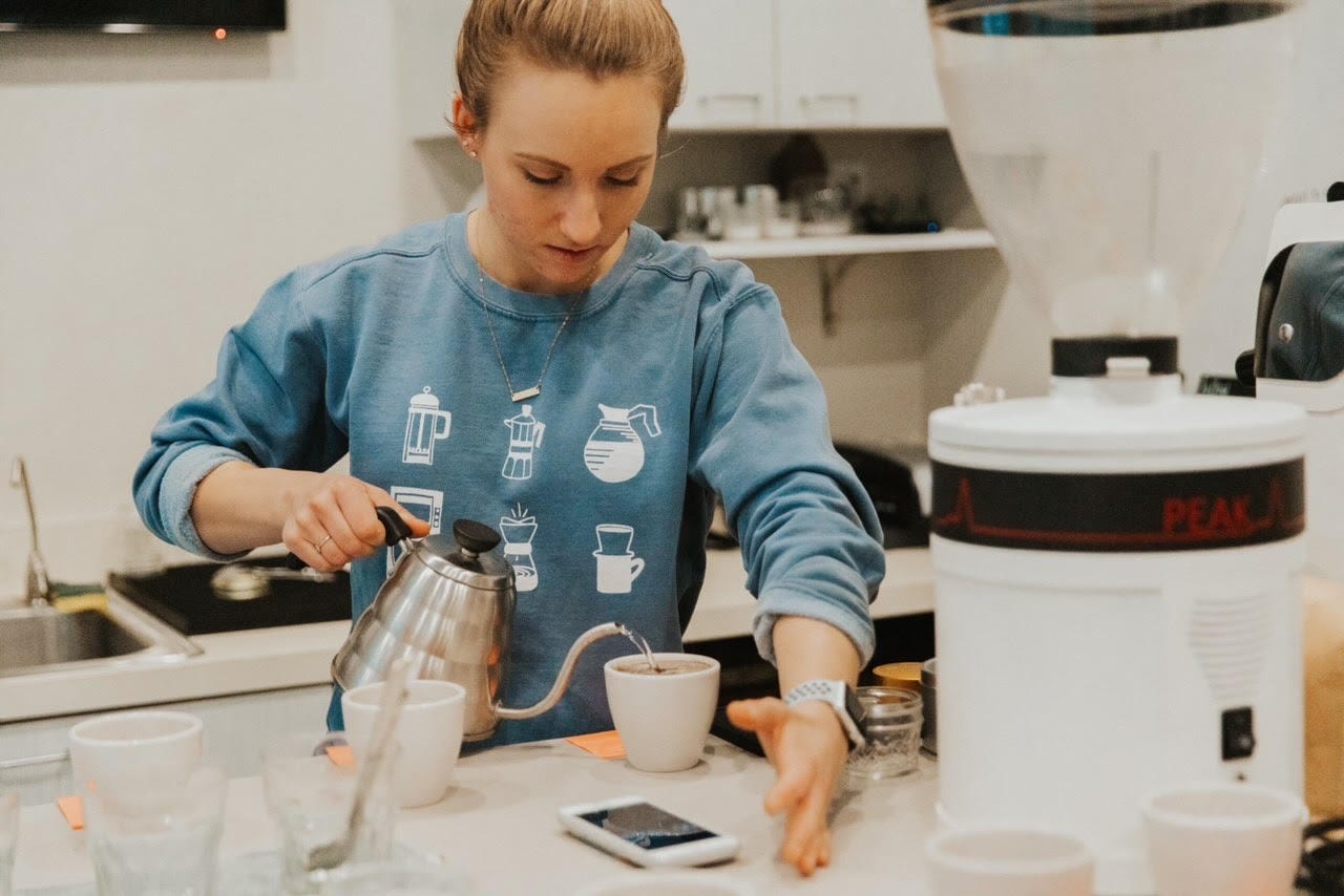 Leap Coffee's head roaster Breanna Briggs in a blue sweatshirt pouring water from silver kettle into a coffee cupping mug in a food laboratory.