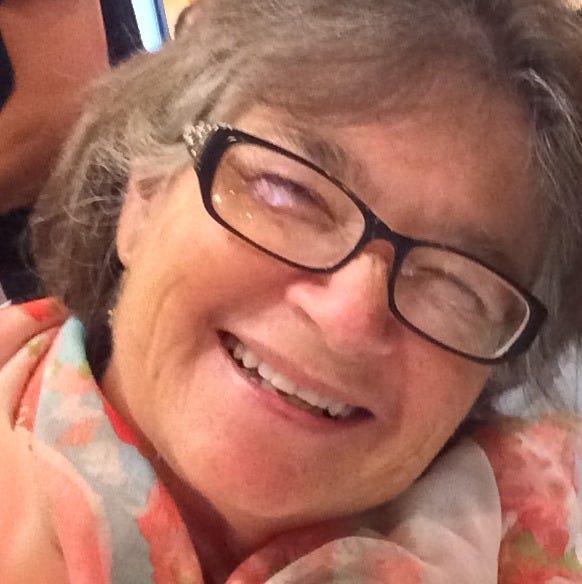 A close up of the author looking stoned - squinty eyes and a vacant smile. The author is white and has on black rimmed glasses and has a scarf in shades of peach around her neck. 