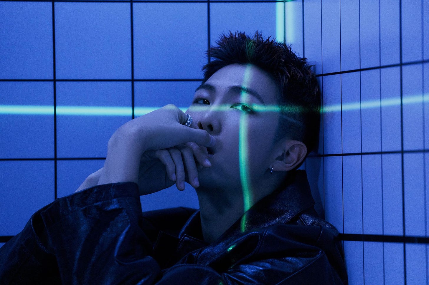 A blue toned picture of Kim Namjoon or RM sitting with a green laser over his eye. He is looking at the camera.