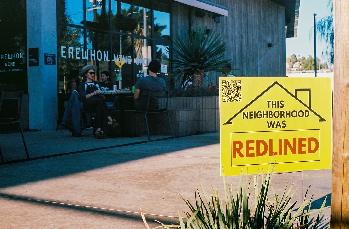 Photograph of the high end grocery store Erewhon in the background with patrons dining at outdoor tables and one of my yard signs in the foreground. 