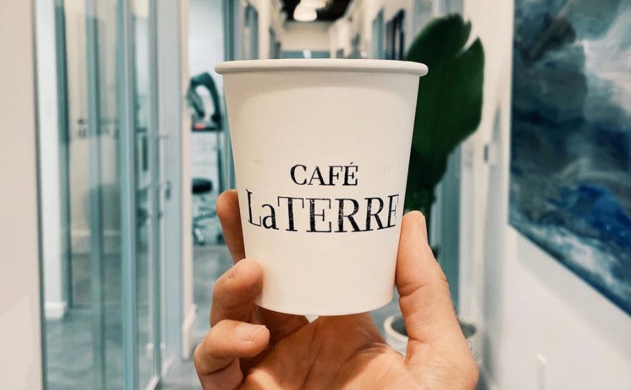 A close up of a white, paper, take out coffee cup with the name Cafe LaTerre inked on it in black. A hand holds the cup right up to the lens of the camera. The cafe's white walls are blurred in the background.