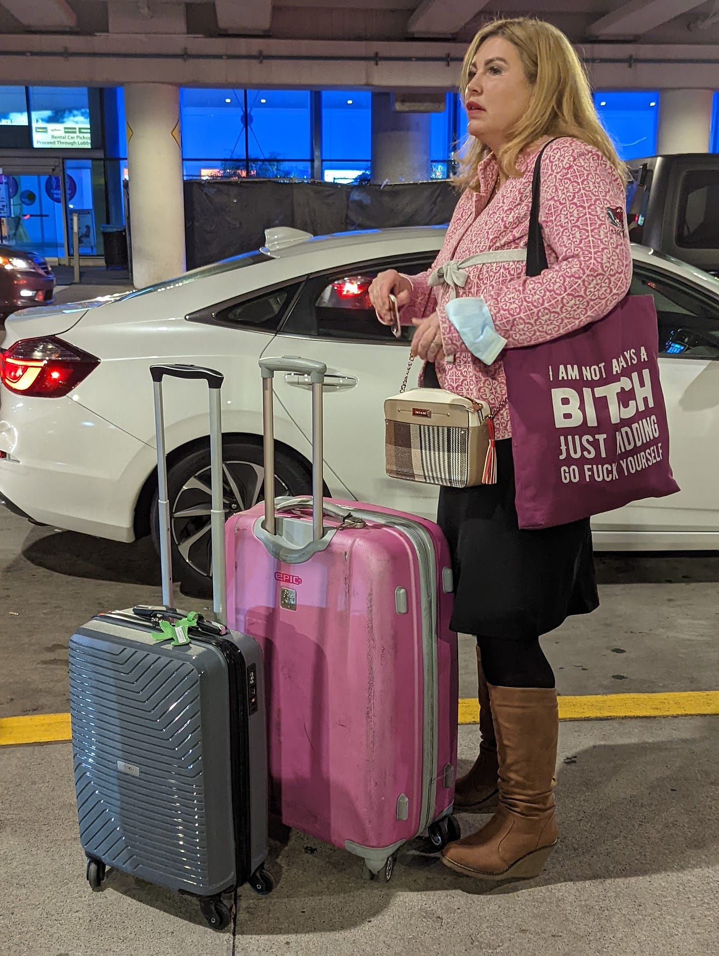 A middle-aged woman in a pink top with a large pink suitcase, carrying a violet tote bag reading: "I'm not always a bitch just kidding go fuck yourself."