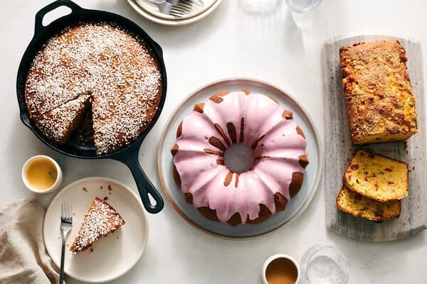A gingerbread cake topped with white pearl sugar is in a cast iron pan with a slice on a white plate next to a cup of coffee. A pink-glazed Bundt cake sits on a platter next to a cheese-topped loaf cake on a cutting board with some slices cut off.
