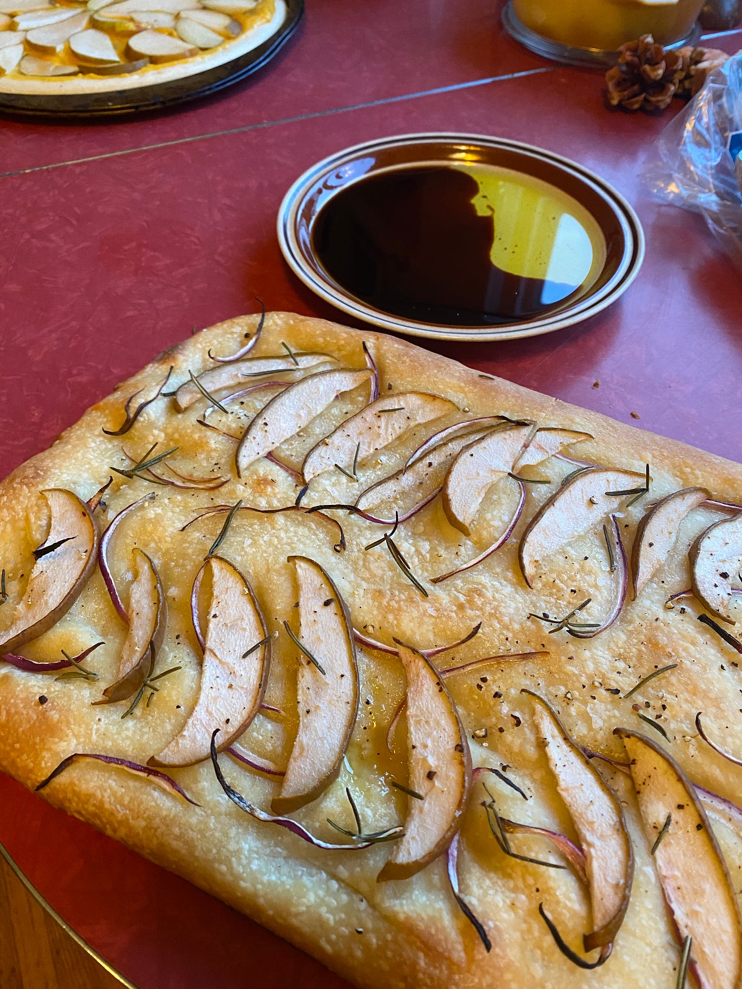 A whole focaccia bread topped with slices of pear and red onion and rosemary leaves on a red table. Behind it is a small plate of oil and balsamic vinegar.