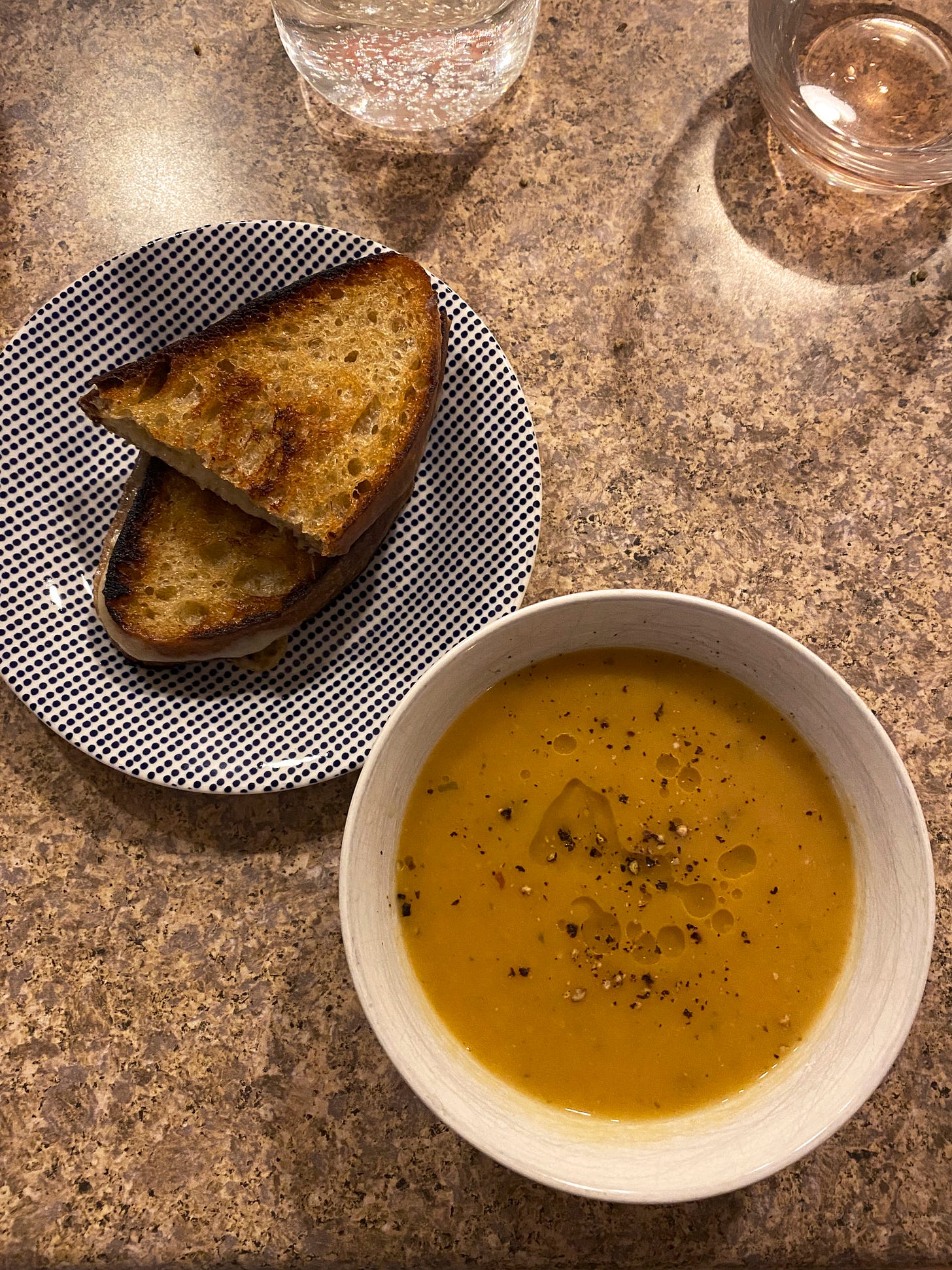 A small white bowl of orange-ish soup, dusted with black pepper and drizzled with olive oil. Slightly above and to its left is a blue and white dotted plate with two halves of a grilled cheese on it. In the right corner is a glass of rosé wine.