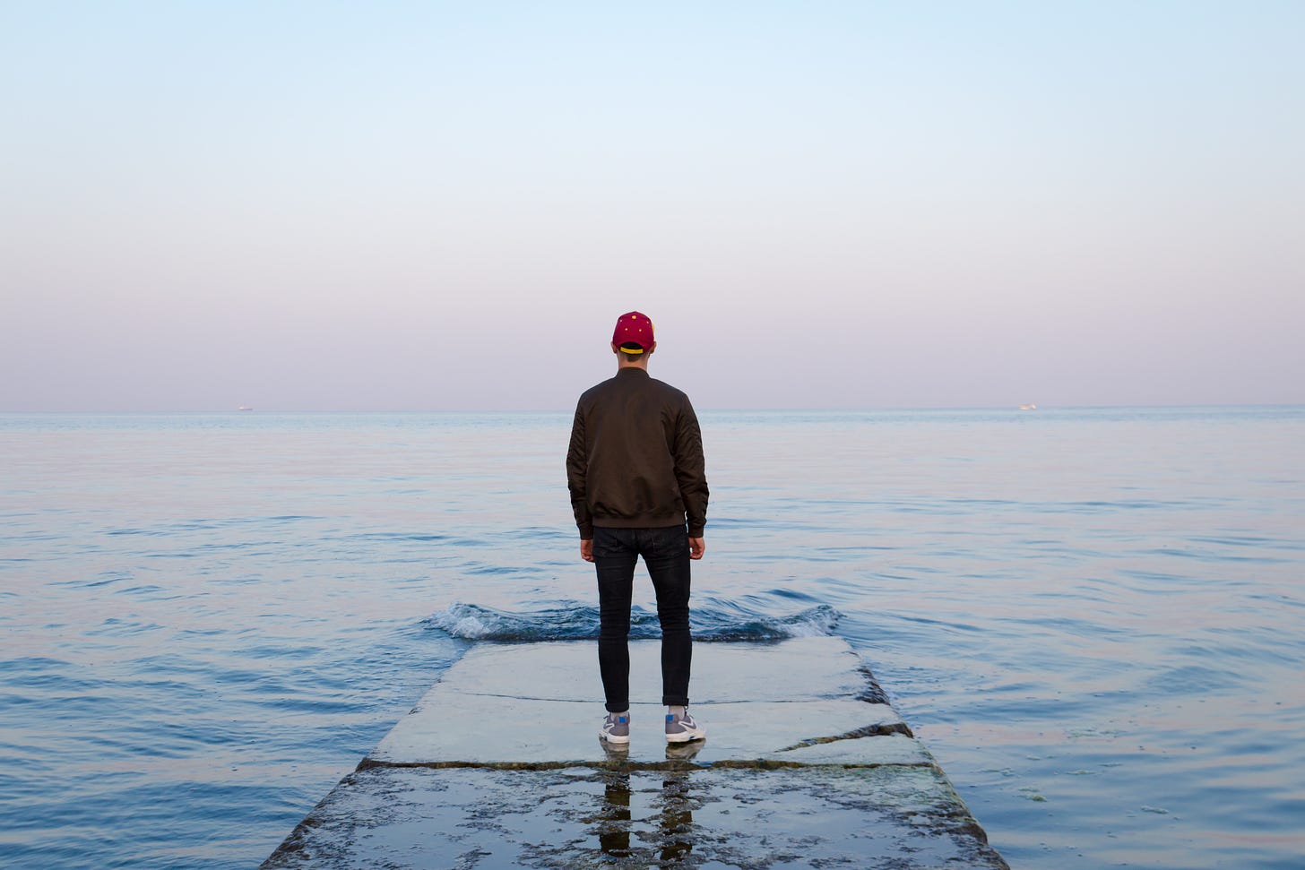 Titelbild: Mit dem Rücken zur Kamera steht ein Mensch in dunkler Kleidung und roter Mütze auf einer Art Steinsteg, der ins Meer (oder einen großen See) führt und kurz vor ihm im Wasser endet. Vor ihm ist eine kleine Welle zu sehen, ansonsten ist die Wasseroberfläche flach. Der Himmel im Hintergrund ist grau.