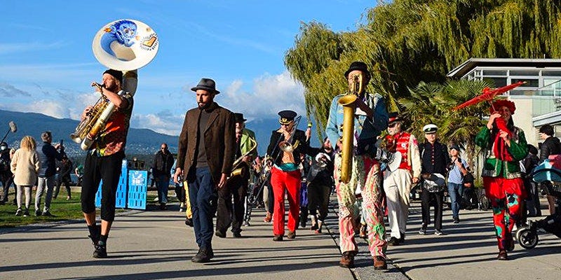 A row of four individuals walk facing frontwards. The central figure wearing a grey fedora hat, a brown jacket and sweater and dark jeans is artist Carmen Papalia. The person on the far left is wearing dark black pants, a colourful shirt, and is playing a large white and bronze tuba that has a drawing of a blue cartoon character coming out of the horn opening. On the right side there is a person wearing pink, green and white patterned pants, a jean jacket and a black hat. They are playing the saxaphone , on the far right is a fourth person who is wearing colourful red, green and black pants, a red, green and black jacket, a red floral flower wreath on their head and they are holding up a red parasol. Behind these four figures are several more rows of people who are all playing different instruments, they all are walking in the same direction as the for figures in the front row. These are all members of the Carnival Band. In the distance behind this band there is a building on the right as well as a large green leafy willow tree. In the background on the left side of the image, there is blue sky spotted with white clouds as well as mountains in the far horizon line. The figures are all walking forwards on a cement pathway flanked by green grass on the left and a building on the right.