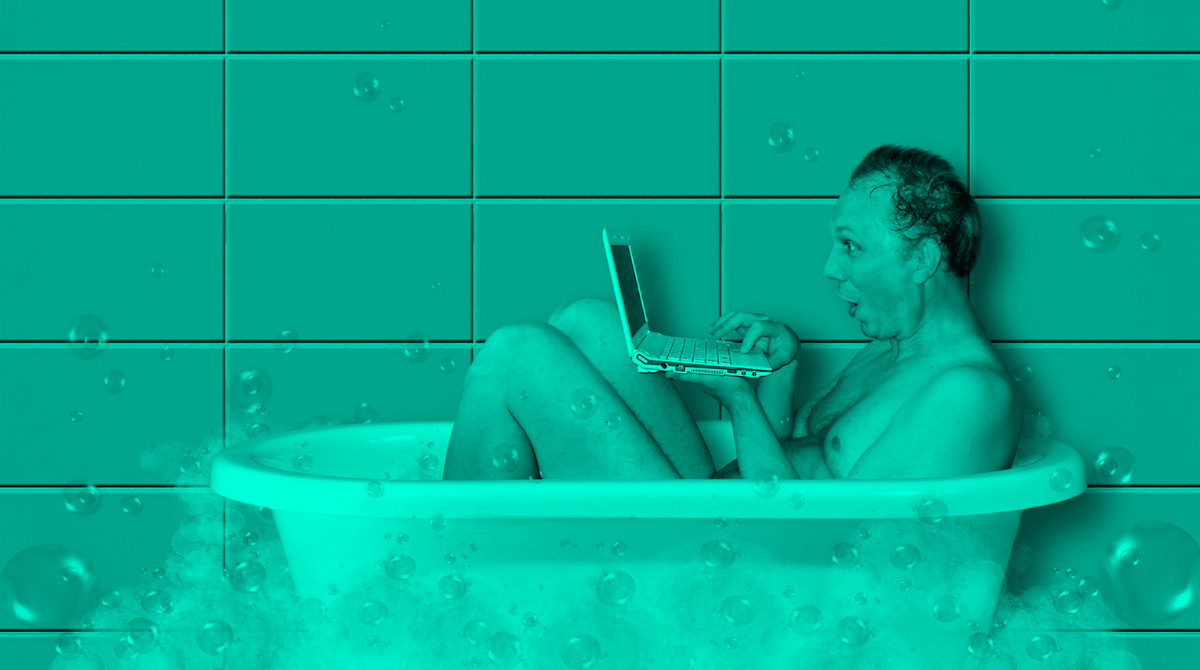 A white man sitting in a bath tub while holding a small laptop. There are bubbles around the outside of the tub.