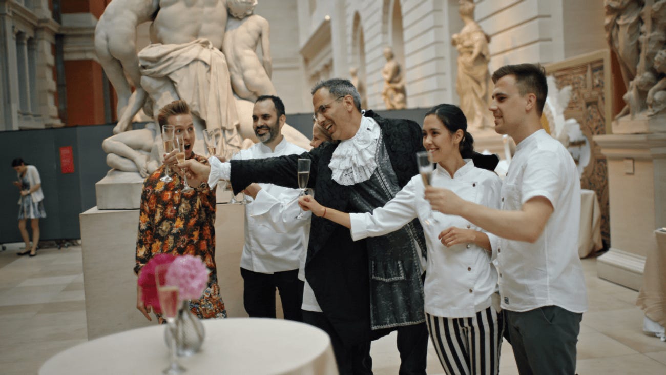 In front of marble statues, Ottolenghi, dressed in an 18th century costume, drinks champagne with four chef colleagues.