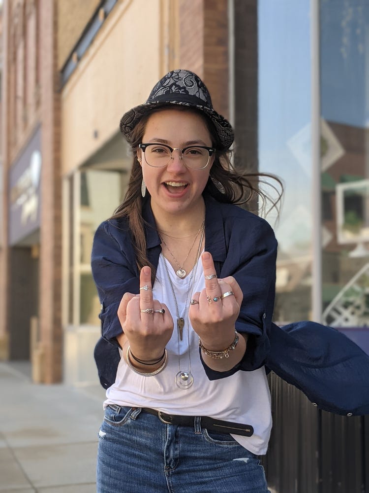 Lydia is standing in a street giving two middle fingers to the camera. She is wearing a hat, a white shirt and blue jacket. She has two rings on each of her middle fingers as well as three necklaces and silver earrings and several bracelets are visible on her arm.