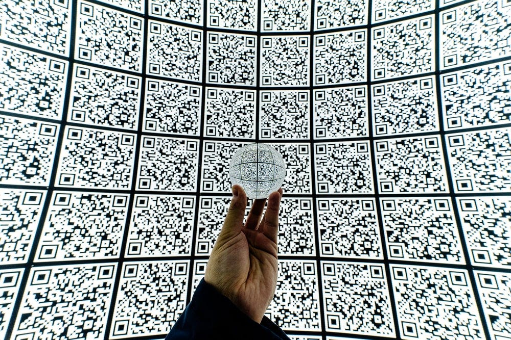 Foto mostra uma mão segurando uma bola de cristal contra um fundo de QR Codes.