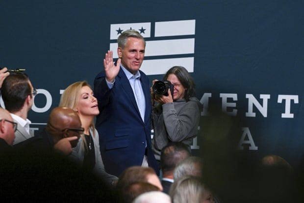 House minority leader Kevin McCarthy waves to people in front of a "Commitment to America" sign as reporters and photographers mill around.