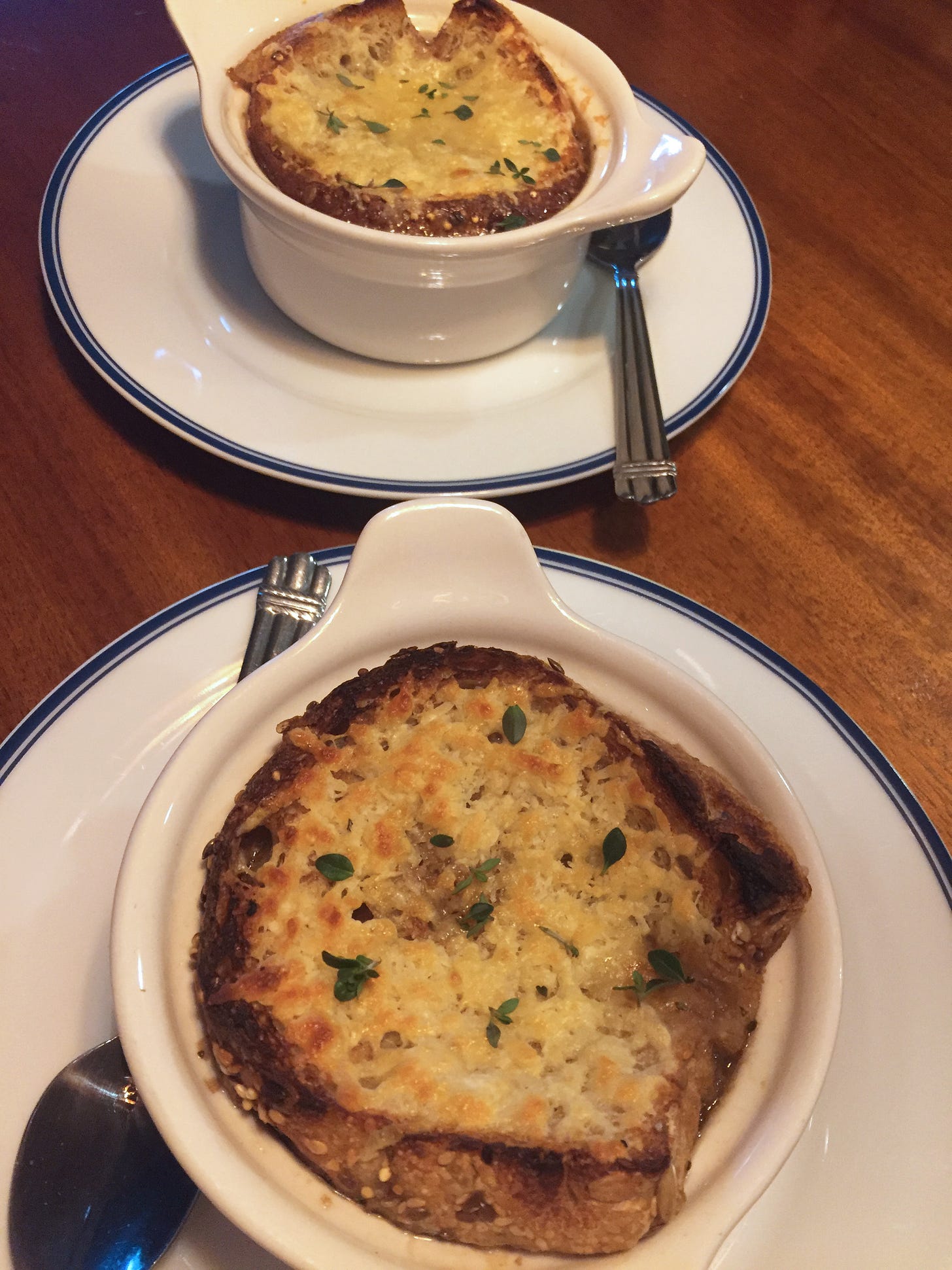 two bowls of French onion soup in traditional white bowls with handles rest on white plates. Browned, melted cheese on toast sits atop the soup.