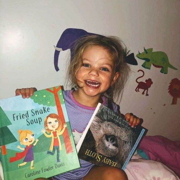 A 5-year old white girl holding an illustrated childrens book titled Fried Snake Soup and another book on sloths, held upside down